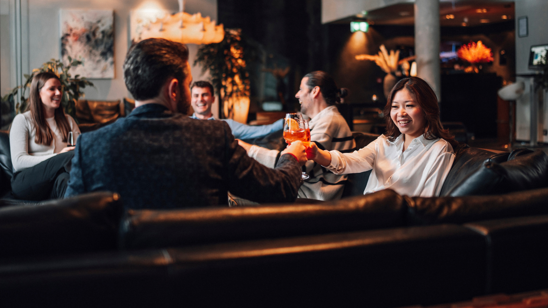  Group of guests enjoying cocktails in the lounge at Thingholt by Center Hotels, seated on black leather sofas surrounded by soft lighting and plants.