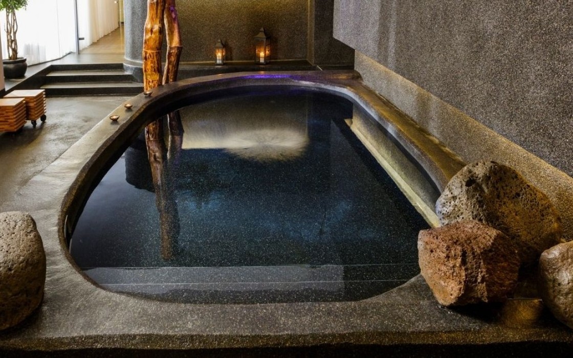 Stone hot tub at Isafold spa inside Thingholt by Center Hotels, surrounded by natural rocks, smooth concrete textures, lantern lighting, and wooden elements that evoke Icelandic nature.