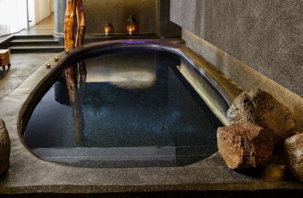 Stone hot tub at Isafold spa inside Thingholt by Center Hotels, surrounded by natural rocks, smooth concrete textures, lantern lighting, and wooden elements that evoke Icelandic nature.