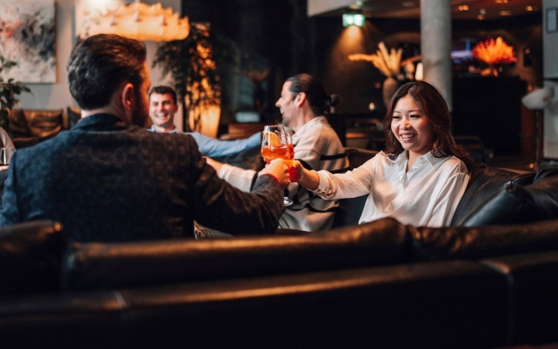 Group of guests enjoying cocktails in the lounge at Thingholt by Center Hotels, seated on black leather sofas surrounded by soft lighting and plants.