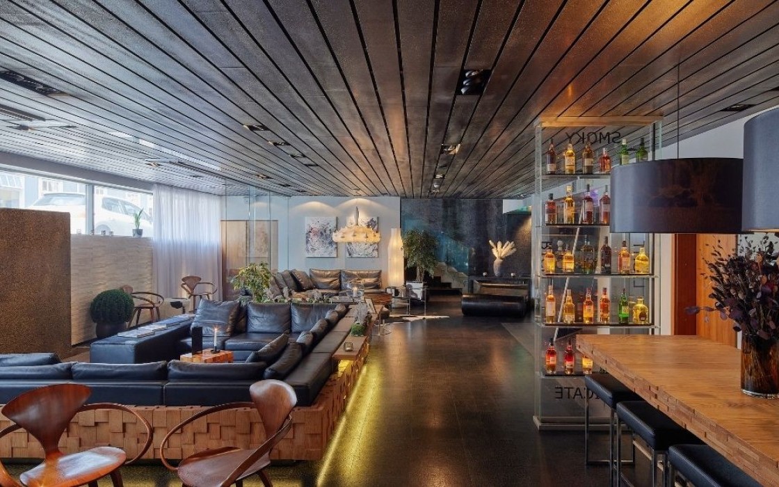 Spacious lounge at Thingholt by Center Hotels featuring dark leather sofas, wooden chairs, glass shelves with spirits, windows and ambient lighting.