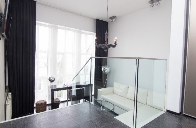 View from the upper level of the Loft Suite at Thingholt by Center Hotels in Reykjavík, showing a white leather lounge sofa, glass railing, black chandelier, tall windows, and a modern writin