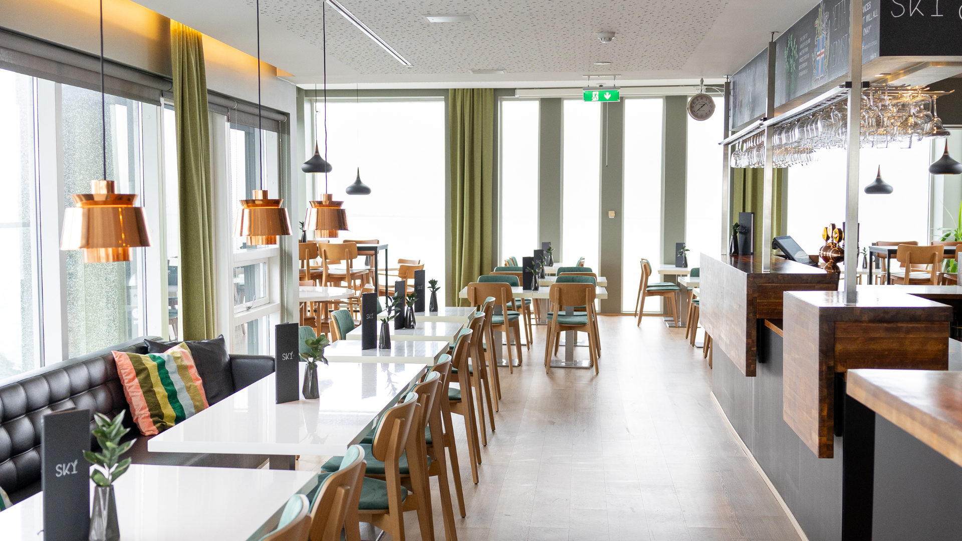 An overview of the top floor bar Sky at Center Hotels Arnarhvoll in Reykjavik with white tables, wooden and green chairs and a dark wood and black bar.