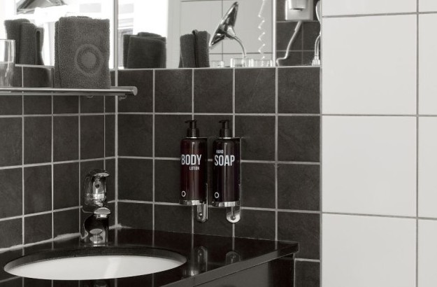 bathroom in the Deluxe Single Room at Center Hotels Arnarhvoll in Reykjavík, featuring dark tiled walls, a sleek black countertop sink, hand soap and lotion dispensers, and folded towels on a