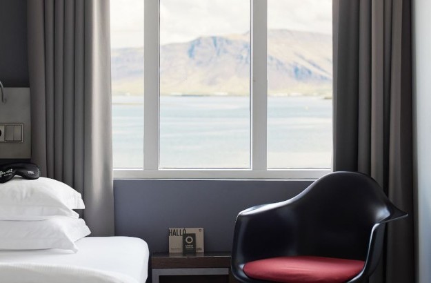 Deluxe Single Room at Center Hotels Arnarhvoll in Reykjavík with a bed positioned in front of large windows, offering a clear view of Faxaflói Bay and Mount Esja, with neutral tones and a bla