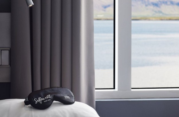 Close-up of a bed in the Deluxe Single Room at Center Hotels Arnarhvoll in Reykjavík, with a black sleep mask resting on white pillows and a serene view of the ocean and mountains through the