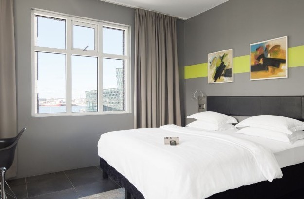 Deluxe Room at Center Hotels Arnarhvoll in Reykjavík, showing the bed with fluffy white bedding, desk, two large windows with city and harbor views, and colorful wall art on gray walls.