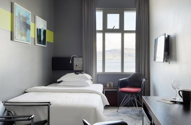 Interior of the Deluxe Single Room at Center Hotels Arnarhvoll in Reykjavík featuring a single bed, wall-mounted TV, abstract art, and panoramic windows showcasing the ocean and mountains.