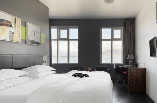 Bright and airy Sea View Room at Center Hotels Arnarhvoll in Reykjavik, featuring a king-size bed, seating area, TV, and panoramic windows offering a scenic ocean and mountain view.