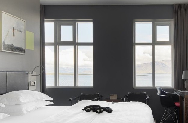 Sea View Room at Center Hotels Arnarhvoll in Reykjavik with a large bed facing two tall windows that overlook the ocean and distant mountains.