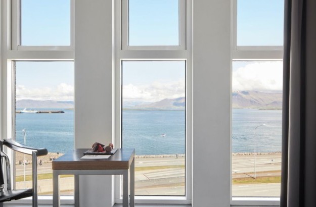 Panoramic view of the ocean and mountains from the Sea View Deluxe Room at Center Hotels Arnarhvoll in Reykjavik, seen through large windows beside a cozy table setup.
