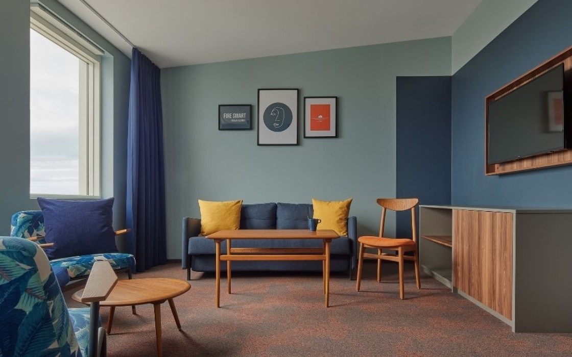 Living room area in the suite at Grandi by Center Hotels in Reykjavik featuring navy sofa, yellow pillows, wall art, and a wall-mounted TV.