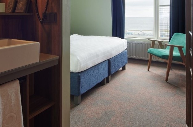 Standard double/twin room at Grandi by Center Hotels viewed from the bathroom area, featuring a bed setup, green chair and wooden finishes.