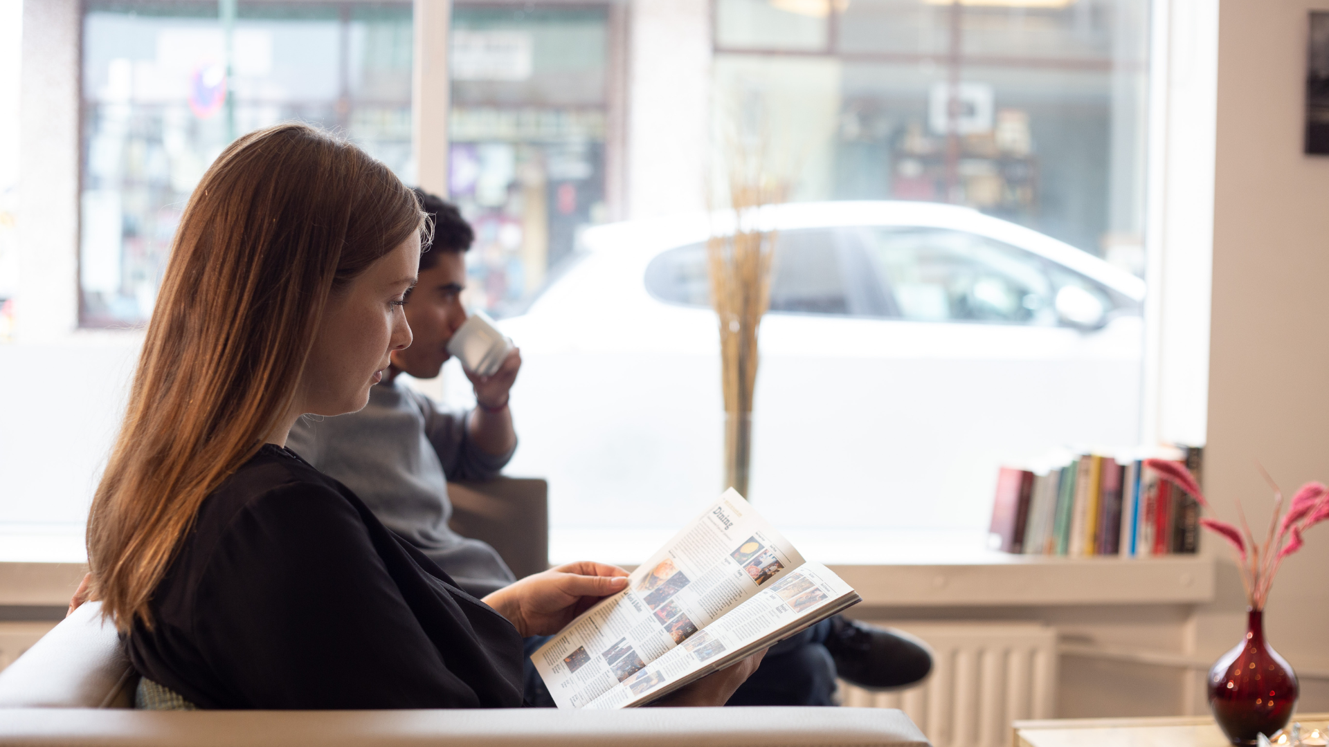 Guest reading a magazine by the window in the lobby lounge at Center Hotels Klopp in Reykjavik, with natural light, street view, and bookshelf in the background.