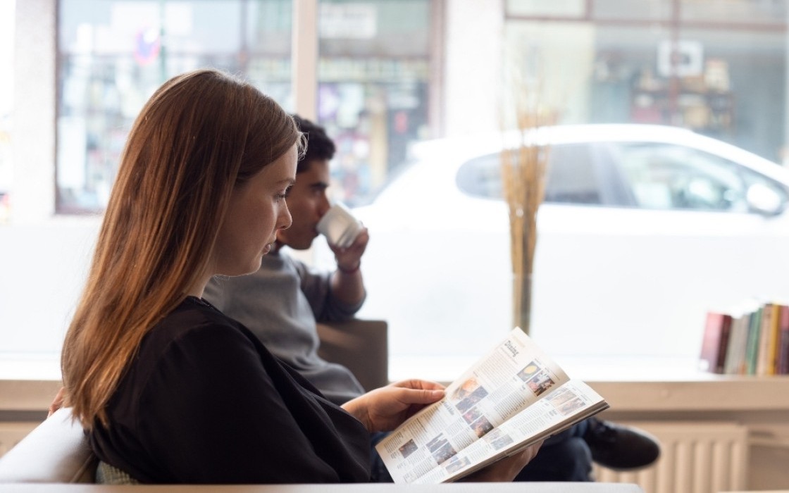 Guest reading a magazine by the window in the lobby lounge at Center Hotels Klopp in Reykjavik, with natural light, street view, and bookshelf in the background.