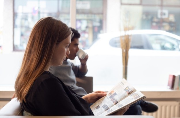 Guest reading a magazine by the window in the lobby lounge at Center Hotels Klopp in Reykjavik, with natural light, street view, and bookshelf in the background.