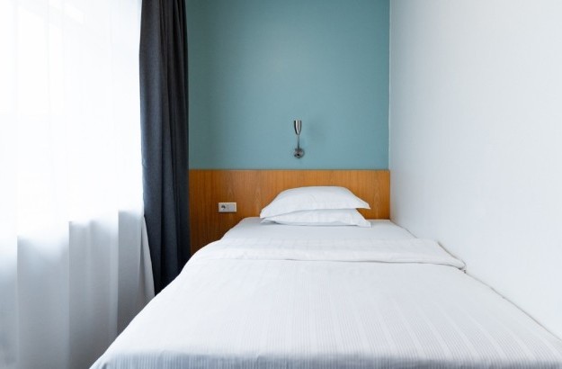 Simple and bright standard single room at Center Hotels Klopp, with a single bed facing a large window, white bedding, and a teal feature wall.