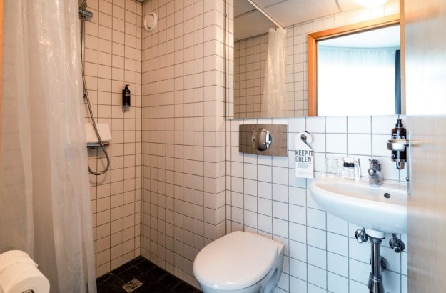 Compact bathroom at Center Hotels Klopp in Reykjavik featuring white tiled walls, a walk-in shower with curtain, wall-mounted soap dispenser, and a white sink and toilet.