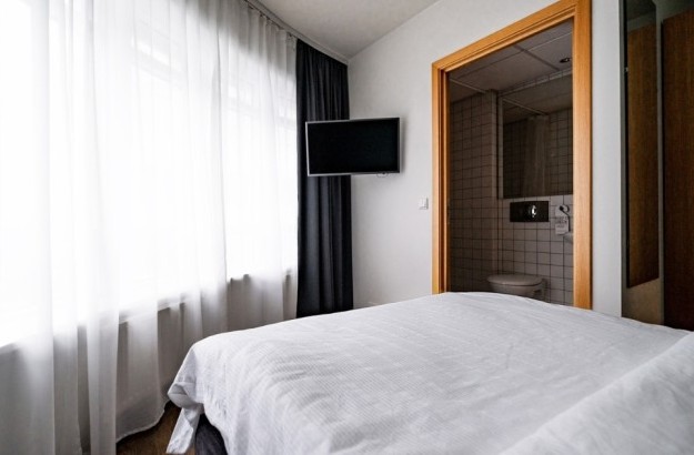 A view from the bed in a superior single room at Center Hotels Klopp. The room features a mounted flat-screen TV, large sheer-curtained windows, and a visible ensuite bathroom entrance.