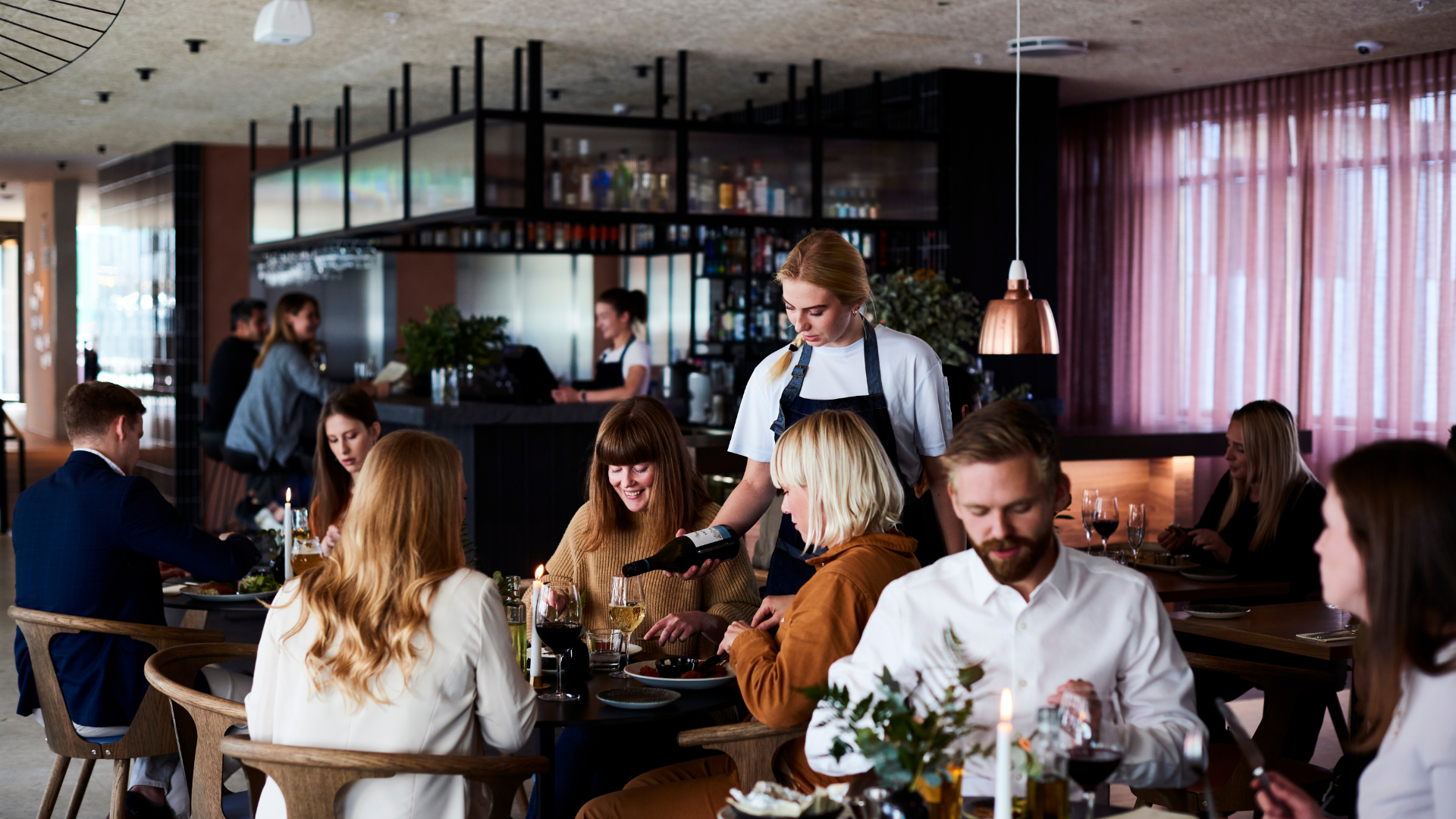 Group of guests dining at Lóa Restaurant at Center Hotels Laugavegur in Reykjavik with wine being poured at a table and bartender in background.