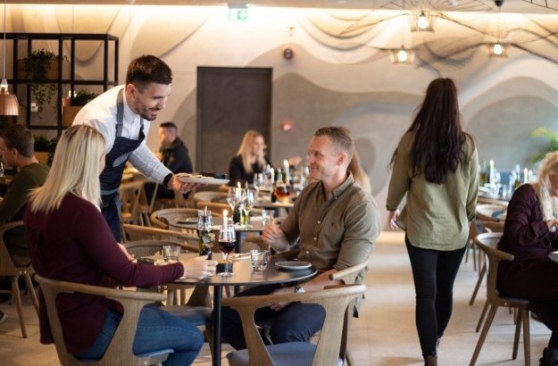  Waiter serving a seated couple in the bright dining area of Lóa Restaurant at Center Hotels Laugavegur in Reykjavik with guests at surrounding tables.