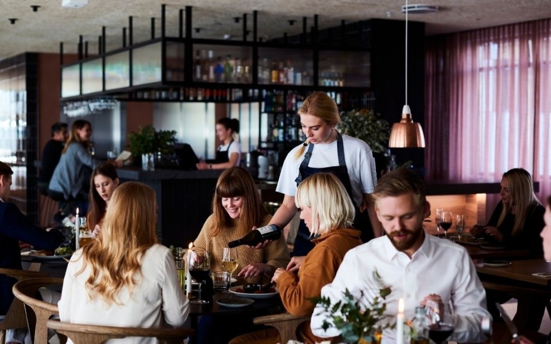 Group of guests dining at Lóa Restaurant at Center Hotels Laugavegur in Reykjavik with wine being poured at a table and bartender in background.