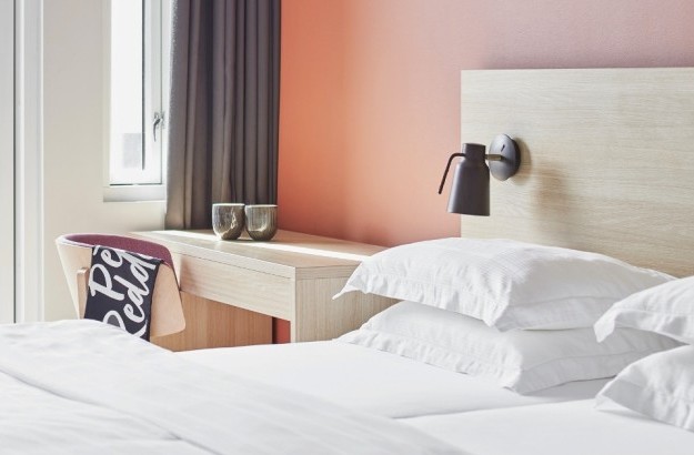 Bed with white linens next to wooden desk and chair, pink wall and blakcout curtains in a deluxe room at Center Hotels Laugavegur in Reykjavik.