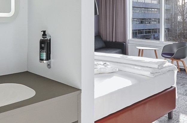 Sink area with mounted soap and partial view of deluxe bed and seating at Midgardur by Center Hotels in Reykjavik.
