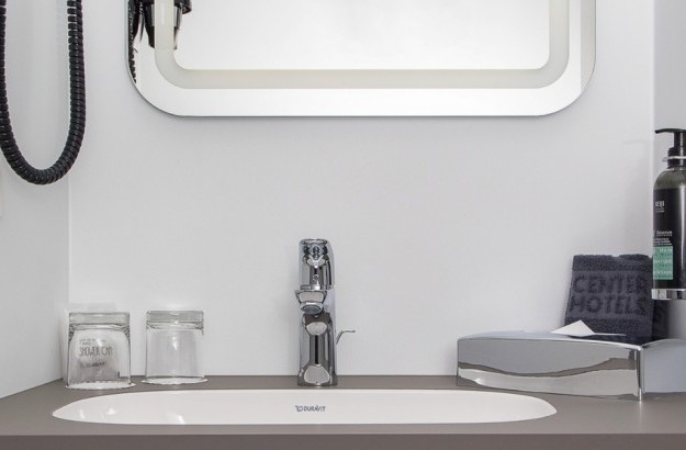 Close-up of a hotel bathroom counter at Midgardur by Center Hotels in Reykjavik, with toiletries, a hairdryer, towels, and a lighted mirror.