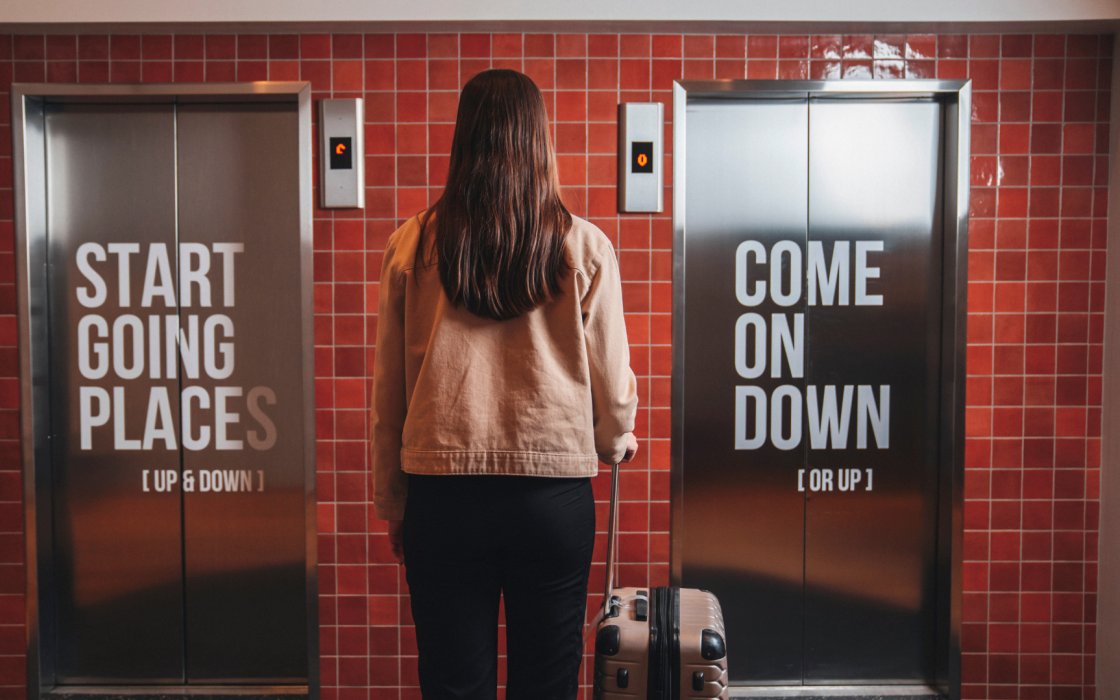 A woman with a suitcase standing between two stainless steel elevator doors at Center Hotels Plaza in Reykjavík. The left door reads “Start Going Places (Up & Down)” and the right says “Come