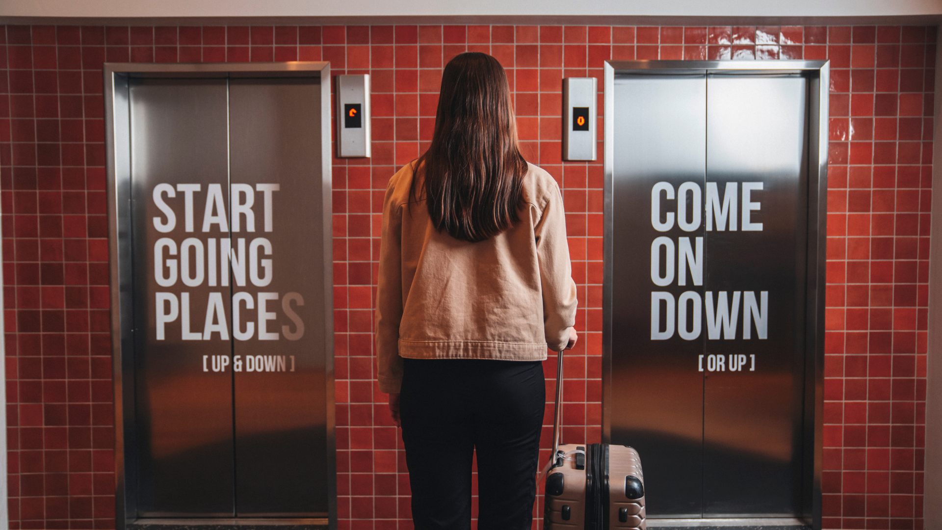 A woman with a suitcase standing between two stainless steel elevator doors at Center Hotels Plaza in Reykjavík. The left door reads “Start Going Places (Up & Down)” and the right says “Come 
