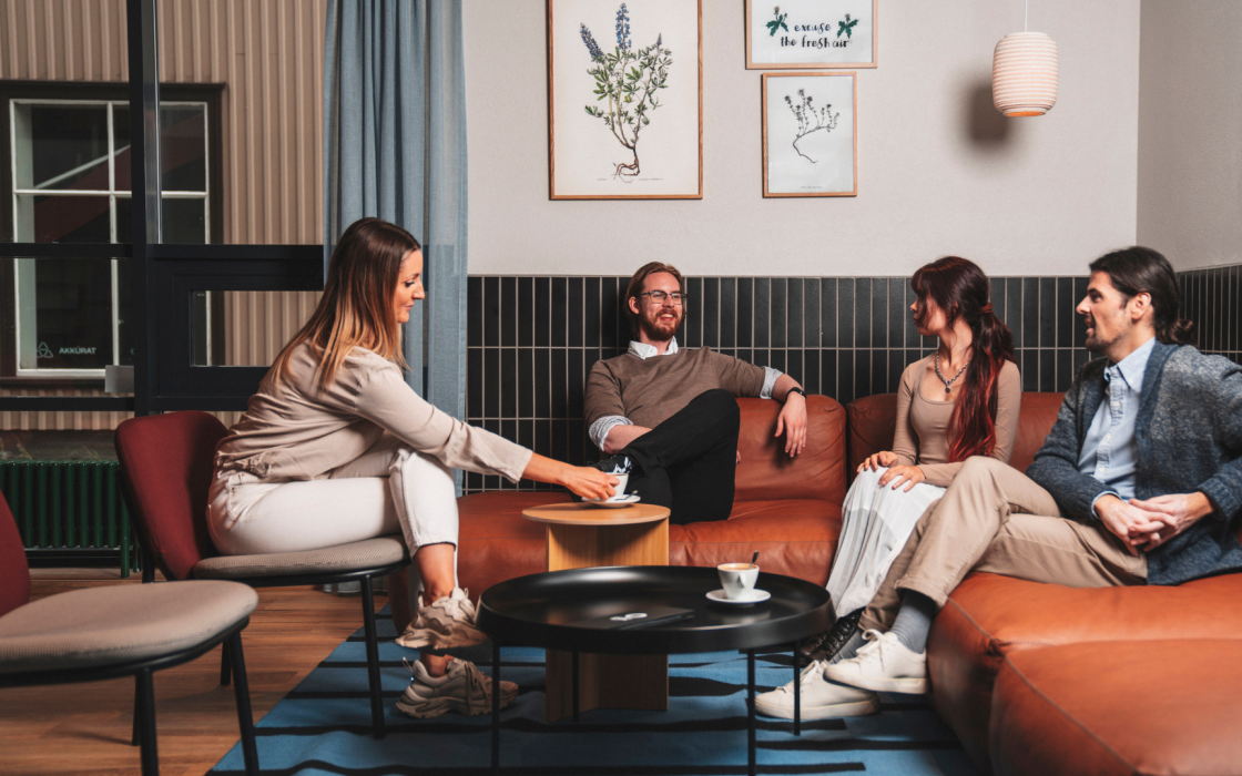 Four people enjoying coffee and conversation in a stylish lounge area at Center Hotels Plaza in Reykjavík. They are seated on a modern leather couch and chairs under botanical-themed wall art