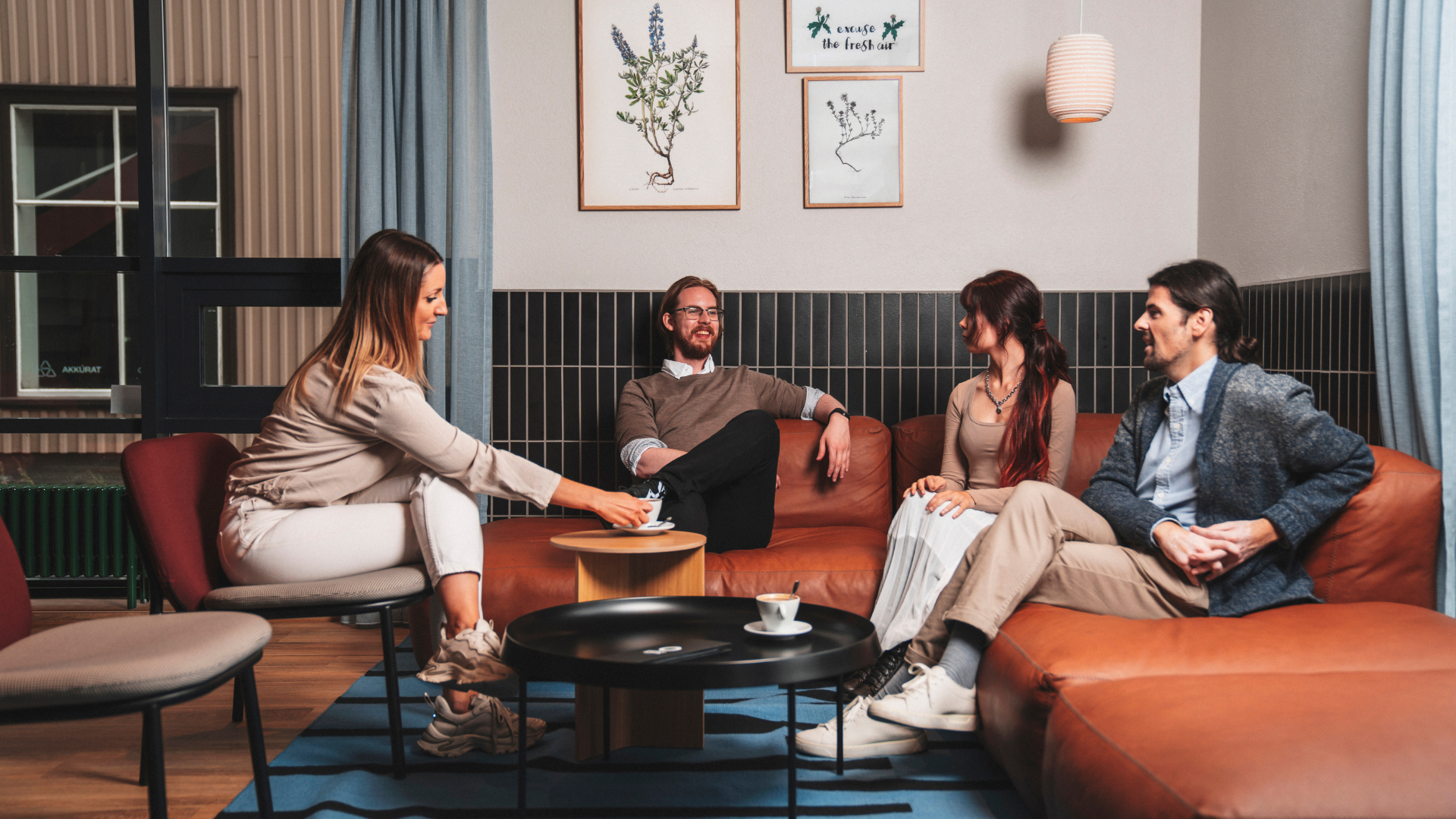 Four people enjoying coffee and conversation in a stylish lounge area at Center Hotels Plaza in Reykjavík. They are seated on a modern leather couch and chairs under botanical-themed wall art