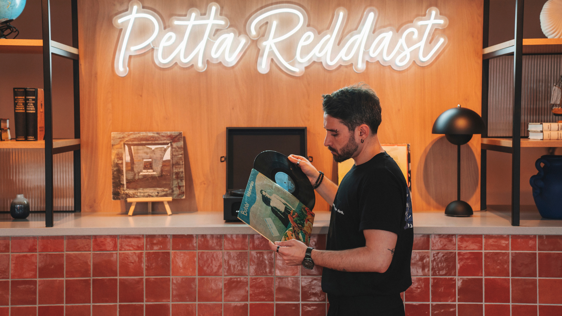 A man flipping through vinyl records in a display area at Center Hotels Plaza in Reykjavík. Behind him is a glowing neon sign that reads “Þetta Reddast,” Icelandic for “It will all work out,”