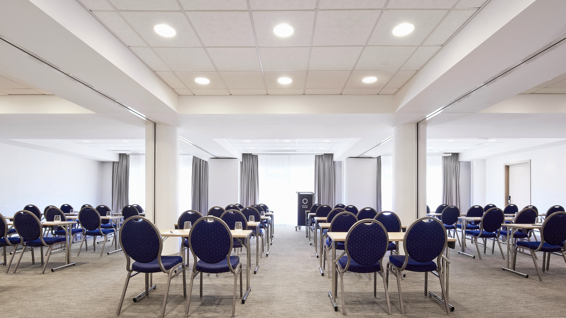 Spacious conference room at Center Hotels Plaza in Reykjavík with rows of blue chairs and desks arranged classroom-style facing a podium. The room is well-lit with ceiling lights and large wi