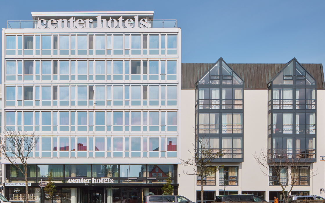 Exterior of Center Hotels Plaza in Reykjavík, showing its modern white facade and adjacent glass-fronted gabled building under a clear blue sky. The hotel entrance is visible at street level