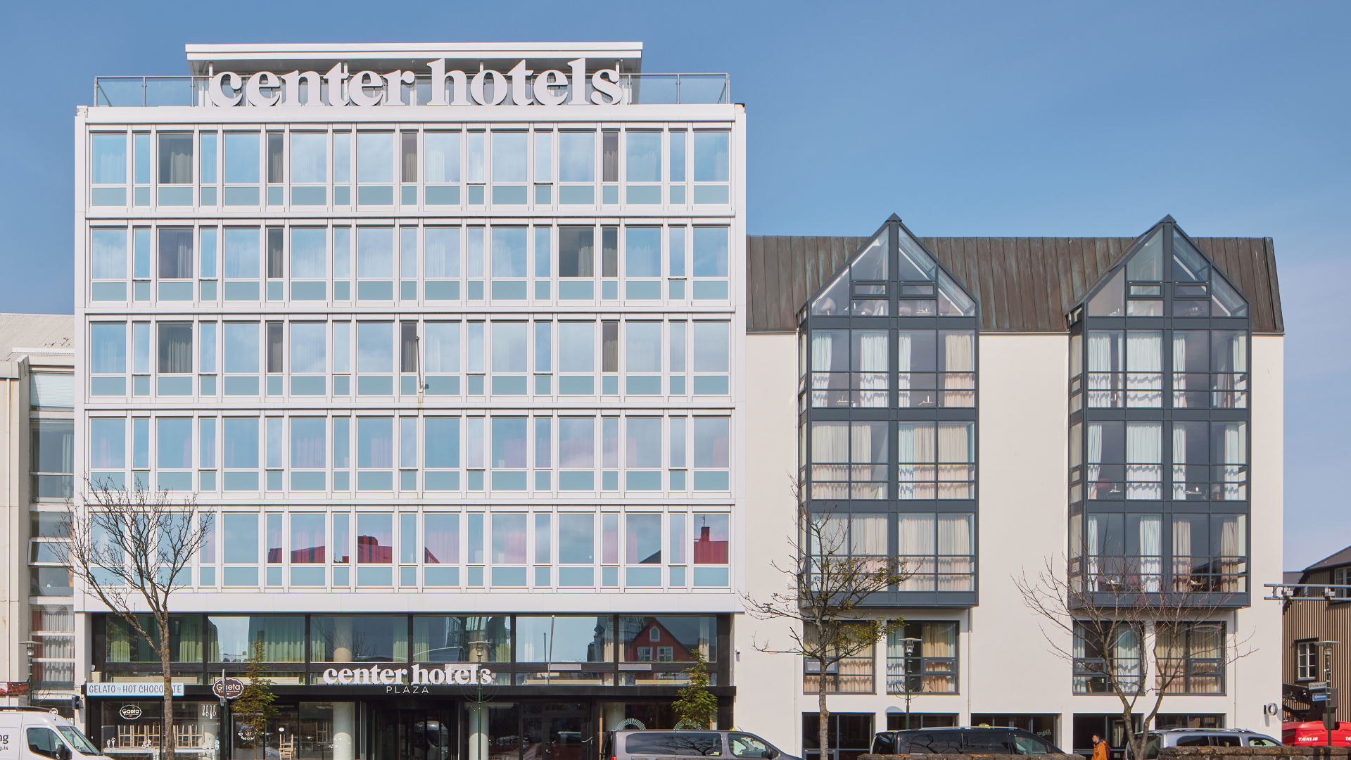 Exterior of Center Hotels Plaza in Reykjavík, showing its modern white facade and adjacent glass-fronted gabled building under a clear blue sky. The hotel entrance is visible at street level 