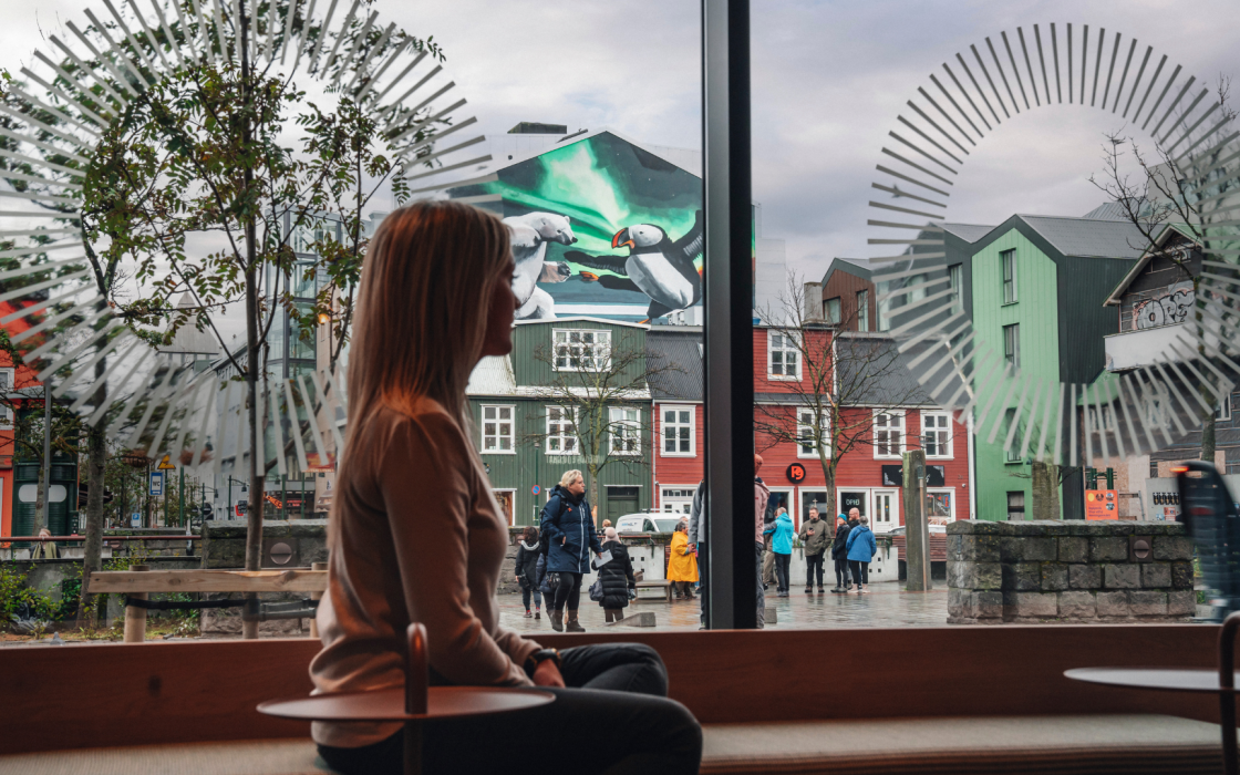 A woman sitting by a large window at Center Hotels Plaza in Reykjavík, looking out at a mural of puffins and polar bears under the northern lights, surrounded by colorful traditional Icelandi