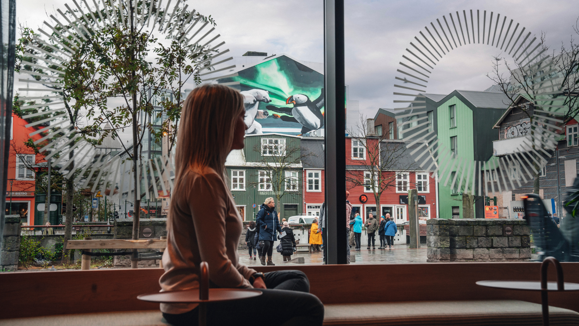 A woman sitting by a large window at Center Hotels Plaza in Reykjavík, looking out at a mural of puffins and polar bears under the northern lights, surrounded by colorful traditional Icelandi