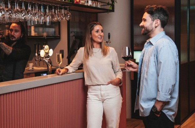 A stylish and intimate moment at the Plaza Bar inside Center Hotels Plaza in Reykjavík, featuring a bartender shaking a cocktail behind a coral-toned counter while a woman and man enjoy a rel