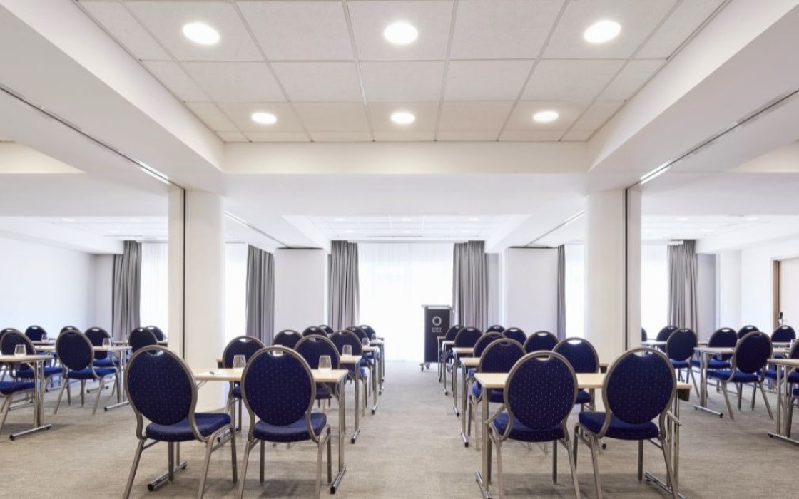 Spacious conference room at Center Hotels Plaza in Reykjavík with rows of blue chairs and desks arranged classroom-style facing a podium. The room is well-lit with ceiling lights and large wi