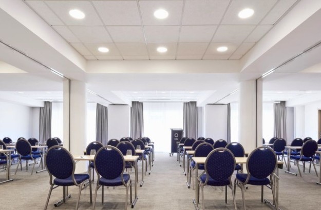 Spacious conference room at Center Hotels Plaza in Reykjavík with rows of blue chairs and desks arranged classroom-style facing a podium. The room is well-lit with ceiling lights and large wi