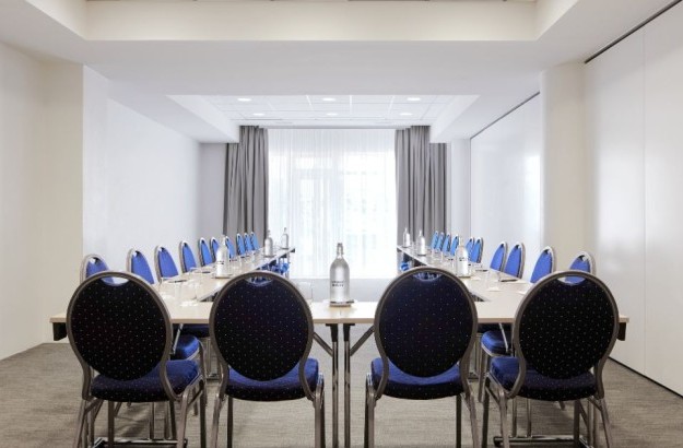 A bright and meeting room at Center Hotels Plaza in Reykjavík arranged in a U-shape setup with navy blue cushioned chairs and minimalist tables. The room features neutral walls, natural light