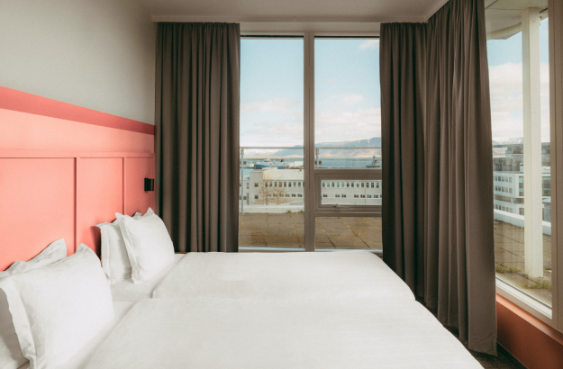 A picture of the bed in the executive room at Center Hotels Plaza with a pink headboard, white linen, and a window with blackout curtain.