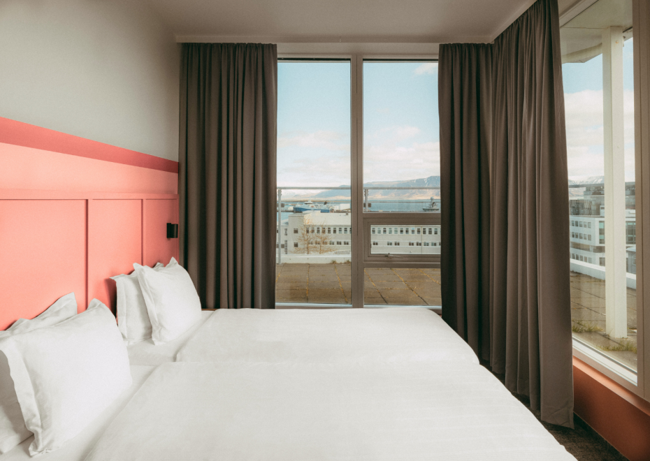 A picture of the bed in the executive room at Center Hotels Plaza with a pink headboard, white linen, and a window with blackout curtain.