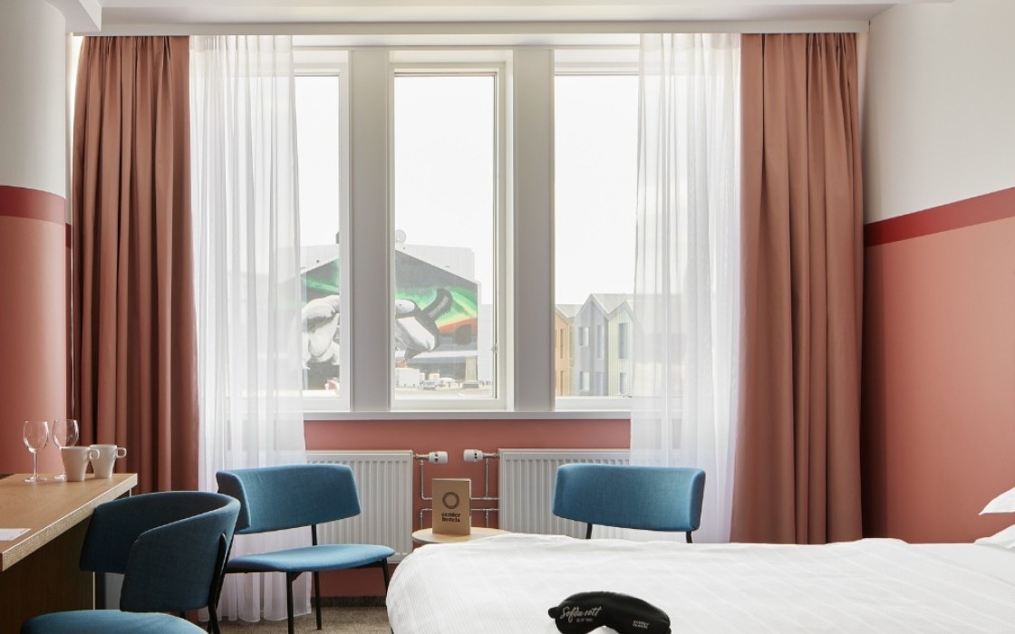 Bright and welcoming Standard Plus Room at Center Hotels Plaza in Reykjavík with a double bed, pastel pink walls, sheer curtains, and blue accent chairs. The room offers a view of downtown Re