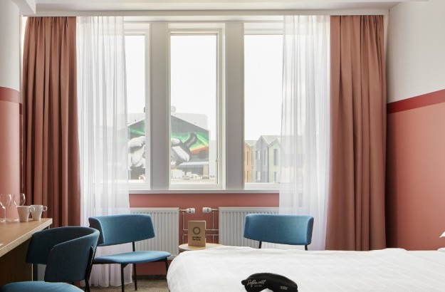 Bright and welcoming Standard Plus Room at Center Hotels Plaza in Reykjavík with a double bed, pastel pink walls, sheer curtains, and blue accent chairs. The room offers a view of downtown Re