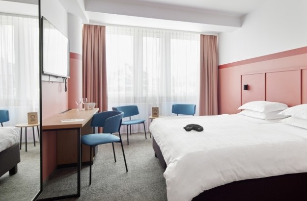 Full view of a Standard Plus Room at Center Hotels Plaza in Reykjavík, showing a neatly made double bed with crisp linens, a writing desk, blue chairs, soft lighting, and large windows with b