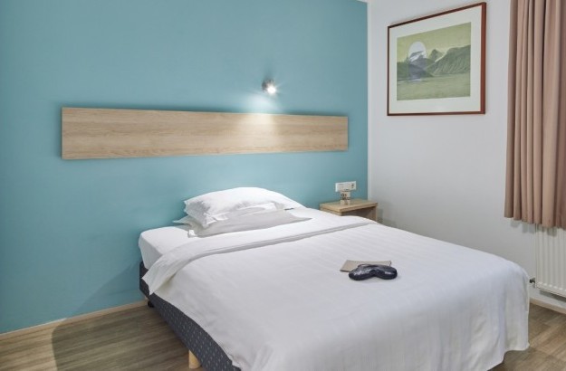 A cozy Standard Single Room at Center Hotels Plaza in Reykjavík with a single bed against a light turquoise accent wall, soft curtains, and natural wood flooring. The space includes framed ar