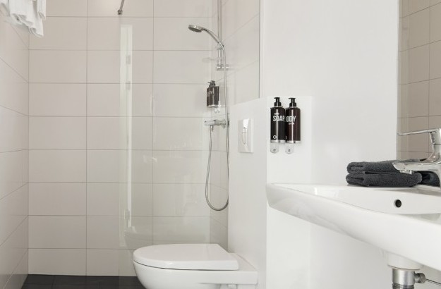 A clean bathroom at Center Hotels Plaza in Reykjavík, featuring white tiled walls, a walk-in shower with mounted dispensers, a sleek wall-mounted toilet, and a compact white sink with dark to